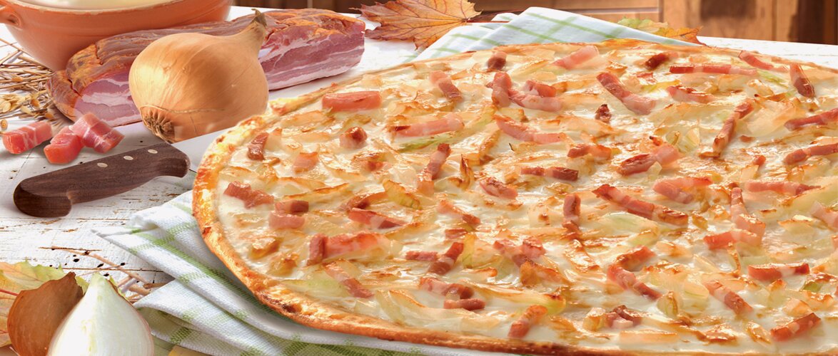 Ein Flammkuchen nach Elsässer Art auf einem grün weiß karierten Tuch dekoriert mit einer Zwiebel, einem Messer, einer Schüssel mit Creme und einer großen Speckscheibe